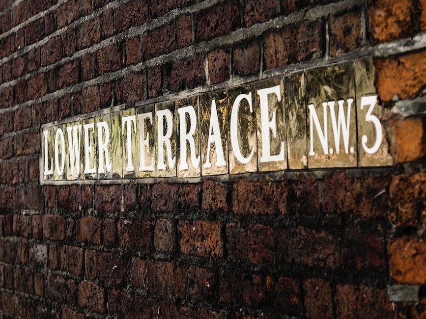 A close-up view of a weathered brick wall featuring a rectangular ceramic street sign with the inscription 'LOWER TERRACE N.W. 3' in bold, white capital letters. The sign has a slightly reflective glazed surface with areas of dirt and moss accumulation, particularly near the edges, indicating exposure to outdoor elements. The textured brickwork behind the sign consists of dark reddish-brown bricks arranged in a traditional running bond pattern, with mortar joints visible between them. The brick surface appears aged and slightly uneven, with some darker patches and subtle variations in colour. The surrounding environment suggests an urban setting, possibly a residential area or alleyway, where the sign is affixed to direct or identify a location. The lighting in the image provides a natural, soft glow that highlights the texture of both the bricks and the ceramic sign, contributing to an overall impression of a static, external street scene, potentially associated with property boundaries or private access points related to waste collection or utility services. The presence of the sign subtly supports the topic of alternative or independent waste management through identifying a specific property or site where rubbish removal services might be required or arranged outside of municipal systems.