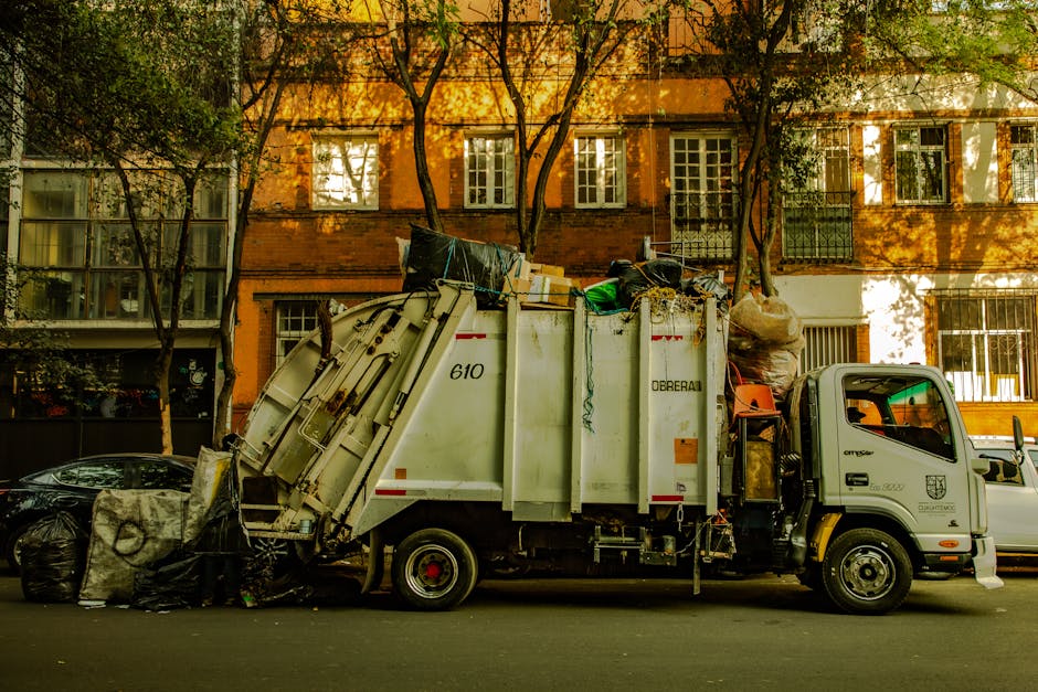 A white waste collection truck with a grey cab is parked on an urban street in front of a multi-storey brick building with several large windows. The truck's rear is loaded with mixed refuse, including black and white bags, cardboard boxes, and various household rubbish. The refuse bins and bags are piled high and slightly overflowing, with some items leaning against the side of the truck, indicating an active rubbish collection or disposal process. The surrounding environment features a row of trees with green foliage casting shadows on the building, which has a warm, amber hue due to sunlight. The street surface is dark asphalt, and part of a silver car is visible on the left, parked adjacent to the truck. An orange container or object is attached or placed near the truck's bulkhead. This scene depicts a typical scene of private waste collection for integrated rubbish removal services, supporting environmentally responsible disposal in an urban setting.