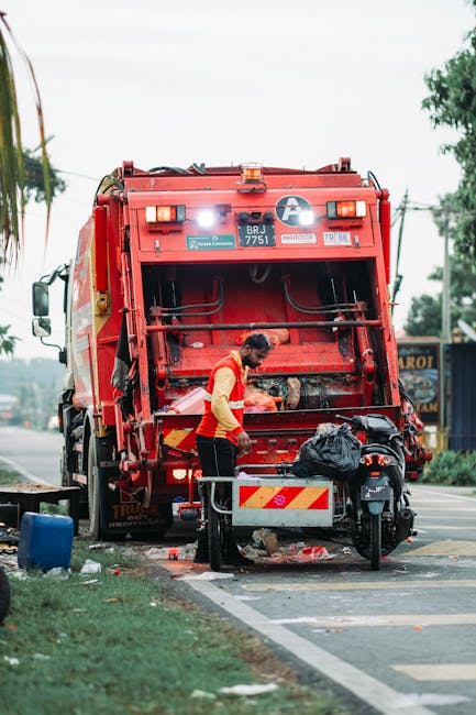 A sanitation worker wearing a high-visibility orange and yellow jacket is engaged in rubbish collection beside a large red refuse collection vehicle parked on the roadside. The rear section of the vehicle, with its open compartment for waste material, displays various mechanical components and safety signals, with the vehicle's identification number 'BRJ 7751' visible on the back. The worker, standing on the pavement, is handling a black rubbish bag, which appears to contain waste, and is positioning it near the collection opening. The environment includes a street scene with trees, utility wires, and distant signage, highlighting a typical urban setting for waste collection services. The surrounding area shows scattered debris and waste, with a small area of grass and the curb edge in the foreground. The sky is overcast, providing diffuse lighting, emphasizing the practical nature of the scene related to private or independent waste disposal activities in compliance with local rubbish collection standards. The precise description supports the context of optimized waste management and alternative rubbish handling services, such as those offered by companies like wastecollectionwesthampstead.co.uk.