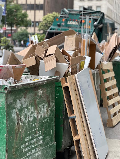 A white waste collection truck with a grey cab is parked on an urban street in front of a multi-storey brick building with several large windows. The truck's rear is loaded with mixed refuse, including black and white bags, cardboard boxes, and various household rubbish. The refuse bins and bags are piled high and slightly overflowing, with some items leaning against the side of the truck, indicating an active rubbish collection or disposal process. The surrounding environment features a row of trees with green foliage casting shadows on the building, which has a warm, amber hue due to sunlight. The street surface is dark asphalt, and part of a silver car is visible on the left, parked adjacent to the truck. An orange container or object is attached or placed near the truck's bulkhead. This scene depicts a typical scene of private waste collection for integrated rubbish removal services, supporting environmentally responsible disposal in an urban setting.