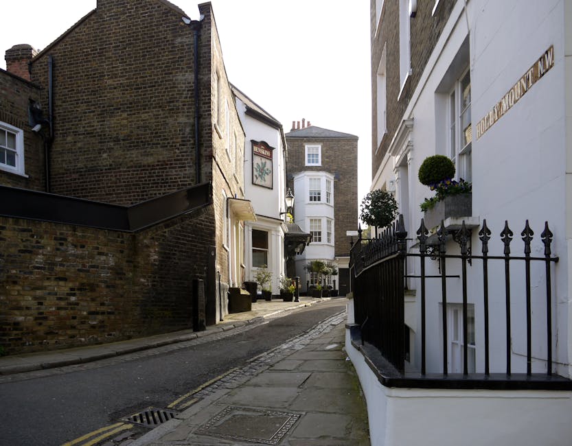 A narrow, paved street in West Hampstead lined with residential buildings, including a white-painted house on the right with decorative black metal railings and potted plants on window sills, and a brick building on the left with a slightly weathered appearance. The street is relatively clean and free of large debris, with a gentle incline leading towards a small, multi-story building at the end of the alley. The sidewalk is built of concrete slabs, with a drain on the street edge, and the overall scene is lit by natural daylight, indicating a clear day. The environment suggests a quiet urban area where private waste collection or on-site rubbish clearance services, such as those offered by wastecollectionwesthampstead.co.uk, might be used to manage local waste and rubbish removal when residents undertake moving or renovation activities, avoiding reliance solely on municipal waste services.