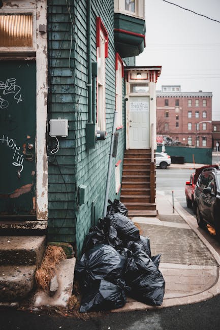 A small urban building with a dark green, weathered wooden exterior and red-trimmed windows. A set of wooden stairs with a white door at the top leads up to an entranceway, situated at the corner of a narrow pavement beside parked vehicles. On the pavement below the stairs, several black plastic rubbish bags are piled, appearing full and tied at the top, likely awaiting collection. To the left, a portion of a metal door or panel with graffiti is visible, along with some chipped concrete steps and a patch of loose soil or debris near the base. The scene is set during daylight with overcast natural lighting, and in the background, the view extends to nearby brick buildings and additional parked cars, reflecting an urban environment possibly associated with private waste collection or waste removal services in West Hampstead. This setting illustrates the common issue of rubbish accumulation outside residential properties, which can be addressed through local or independent waste collection options, supported by companies like wastecollectionwesthampstead.co.uk facilitating rubbish removal for tenants and homeowners alike.
