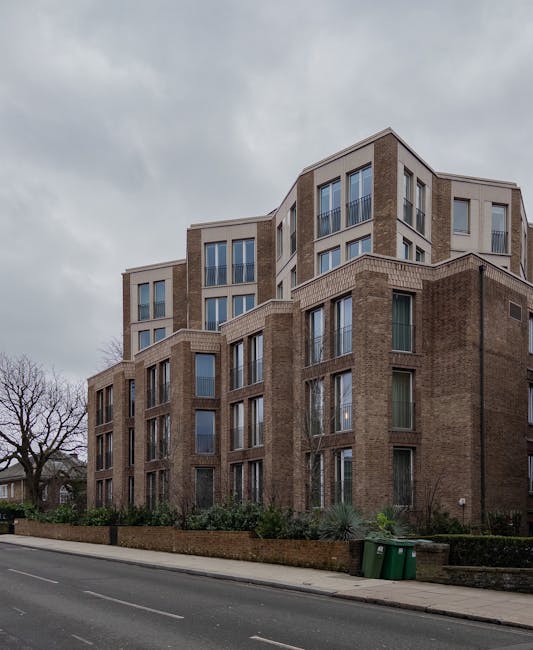 A multi-storey residential building constructed with light brown brickwork featuring large, vertically oriented windows with metal frames; some windows have glass balcony railings. The building has a modern, tiered design with protruding sections at various levels, creating a layered appearance. In front of the building, there is a low brick boundary wall with some greenery and small shrubs growing behind it. On the pavement adjacent to the wall, two green waste bins are positioned next to each other. The street in the foreground is empty, with a smooth asphalt surface marked by white dashed lines. Overcast sky with grey clouds provides diffuse lighting, typical of a cloudy day. The environment suggests a quiet urban neighbourhood where private waste collection or on-site clearance might be employed, as indicated by the presence of waste bins close to the property, reflecting an alternative to regular council rubbish collection for residents or property management, aligned with the context of private rubbish removal services such as those offered by wastecollectionwesthampstead.co.uk.