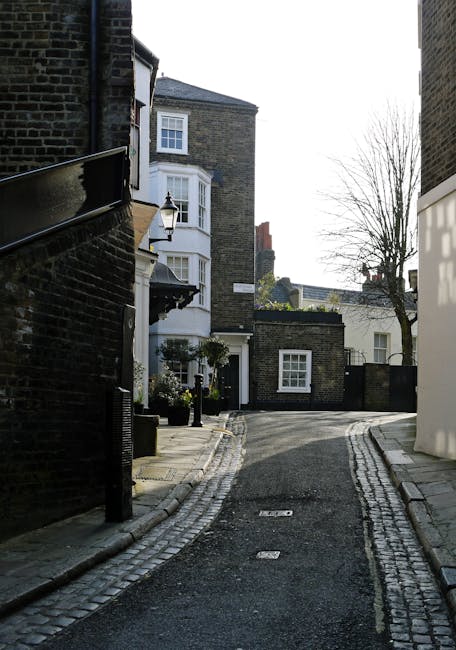 A narrow residential street in West Hampstead, showing a small, winding tarmac road bordered by cobblestone edges on both sides. On the left, there is a dark brick building with a black metal railing and a traditional outdoor lamp attached to the wall. Adjacent, a building with a white bay window featuring white framing and a small, decorative ledge beneath it. Further along, a white door is partially visible, surrounded by potted plants and a small black fence. On the right, a light-colored modern building with smooth external surfaces contrasts with the older brick structures. In the background, there are leafless trees and additional townhouses with flat or sloped roofs, indicating a typical urban neighbourhood. The scene is lit by natural daylight, casting subtle shadows, with the overall environment appearing quiet and residential, evoking a typical setting for alternative waste disposal or private rubbish collection services in West Hampstead, with a focus on local urban street scenes.