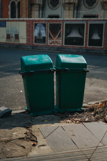 Two green plastic wheelie bins positioned next to each other on a paved sidewalk at the edge of a street, with their lids closed. The bins are made of sturdy plastic with textured surfaces and are slightly weathered, showing minor scuffs and marks. They are situated on a concrete curb with some fallen leaves and small debris scattered around their base. Behind the bins, there is an asphalt road extending into the background, with a brick building featuring decorative architecture and multiple windows in the distance. The scene is illuminated by natural daylight, casting soft shadows on the ground, and the overall environment suggests a typical urban setting suitable for private rubbish collection and waste management services that offer alternatives to traditional council clearance, as might be discussed in the West Hampstead West End Lane rubbish collection guide.