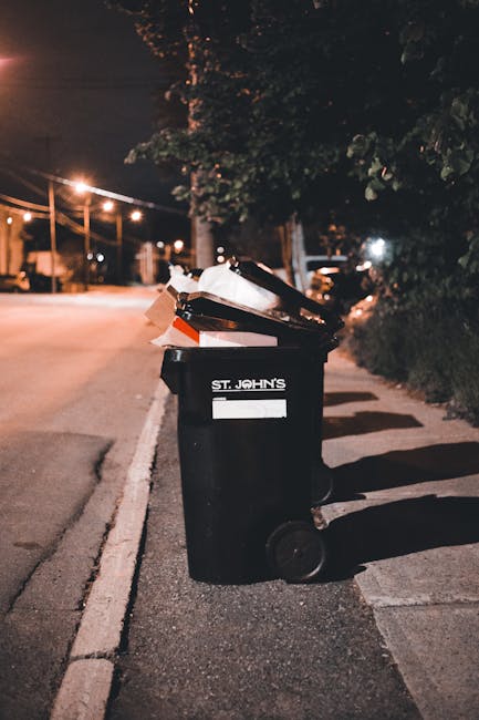 A black wheelie bin with the label 'ST. JOHN'S' is positioned on the pavement next to a curb in a residential area during nighttime, with the bin's lid open revealing an overflow of waste materials, including cardboard boxes and plastic packaging. The bin is made of sturdy plastic, with a textured surface and two large wheels at the base for mobility. It stands in front of a hedge and trees that cast shadows on the ground, and a few streetlights illuminate the scene, creating a warm, low-light atmosphere. The surrounding environment features a quiet street with visible road markings, and additional bins or waste containers are partially visible further down the pavement. The scene reflects urban waste management practices, possibly involving independent rubbish collection, emphasizing the importance of proper waste disposal and clean streets, which wastecollectionwesthampstead.co.uk services support through alternative rubbish handling solutions, including same-day collection options.
