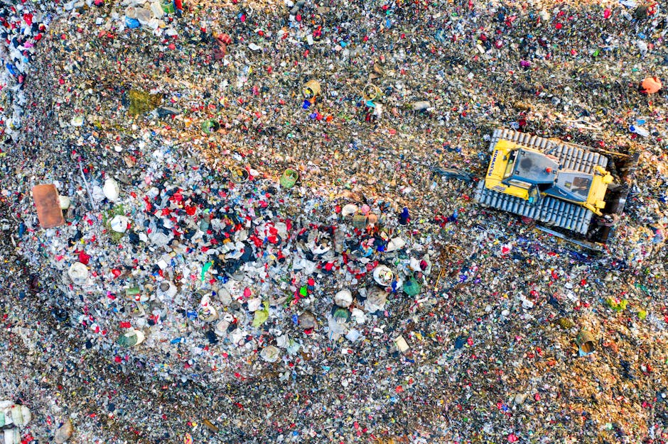 An aerial view of a large landfill site with a yellow bulldozer equipped with a tracked undercarriage and covered blade, situated on the right side of the image. The site is densely packed with various types of waste materials, including plastic, paper, and miscellaneous debris, mostly in shades of white, red, blue, and green, creating a textured, cluttered surface. The waste covers a significant expanse, with some areas appearing more compacted than others, and the bulldozer appears to be actively managing or leveling the refuse. The environment around the waste is exposed soil, with a mixture of rough, uneven textures and some visible tire tracks from the machinery. The lighting suggests natural daylight, casting slight shadows on the uneven terrain and emphasizing the chaotic, mixed nature of the refuse. This scene illustrates the scale of modern waste disposal and highlights the importance of professional rubbish removal services like those offered by wastecollectionwesthampstead.co.uk, particularly for managing large volumes of waste in an organized, efficient manner with an emphasis on proper site handling and environmental considerations.