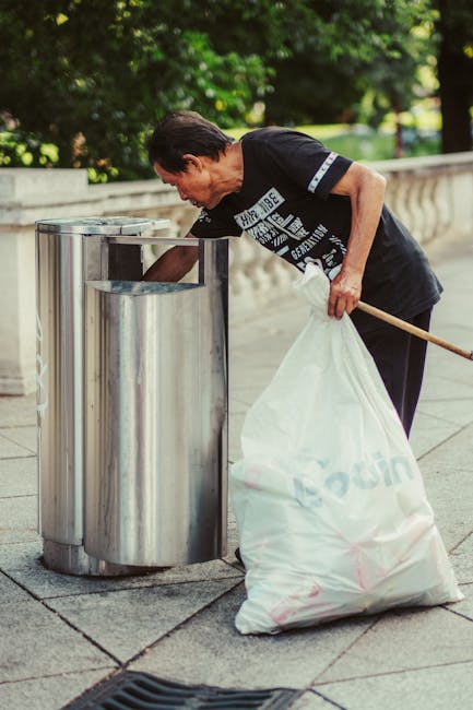 A black wheelie bin with the label 'ST. JOHN'S' is positioned on the pavement next to a curb in a residential area during nighttime, with the bin's lid open revealing an overflow of waste materials, including cardboard boxes and plastic packaging. The bin is made of sturdy plastic, with a textured surface and two large wheels at the base for mobility. It stands in front of a hedge and trees that cast shadows on the ground, and a few streetlights illuminate the scene, creating a warm, low-light atmosphere. The surrounding environment features a quiet street with visible road markings, and additional bins or waste containers are partially visible further down the pavement. The scene reflects urban waste management practices, possibly involving independent rubbish collection, emphasizing the importance of proper waste disposal and clean streets, which wastecollectionwesthampstead.co.uk services support through alternative rubbish handling solutions, including same-day collection options.